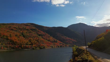 Drone-up-over-lake-reservoir-looking-at-mountains-covered-in-fall-autumn-October-November-trees-changing-color-to-orange-red-and-yellow,-panning-over-to-road-and-backing-up-beautiful-leans-flair