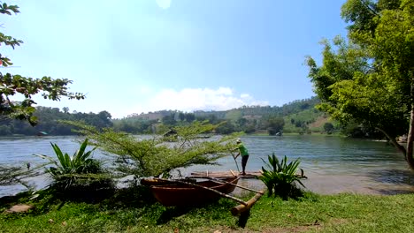 Mann-Auf-Einem-Bambusfloß-Im-See-Mit-Nahaufnahme-Eines-Kleinen-Bootes-Und-Grünem-Gras-An-Der-Vorderseite-Sowie-Horizont-Und-Blauem-Himmel-Im-Hintergrund