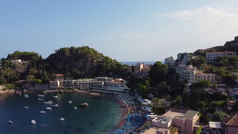 Drone-shot-revealing-a-beautiful,-tranquil-bay-in-Taormina,-Sicily-with-resorts,-hotels,-apartments-on-hills,-cypress-trees,-blue-sea,-boats,-umbrellas-and-sunbeds-on-the-beach