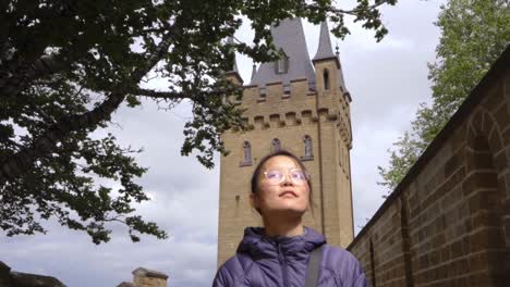 An-awe-inspired-tourist-admires-the-grandeur-of-Hohenzollern-Castle,-a-historic-German-fortress,-set-against-a-lush-backdrop-along-old-brick-wall-pathway