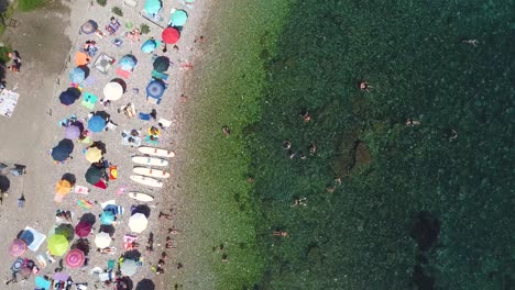 Pan-over-aerial-drone-shot-of-a-pebble-beach-in-Taormina,-Sicily-in-Italy-with-people-swimming,-relaxing-and-suntanning