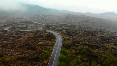 AERIAL:-Drone-following-a-car-driving-down-the-road-weaving-around-dark-lava-rocks-of-active-volcano-Mount-Etna,-Sicily-Italy,-revealing-unique-landscape