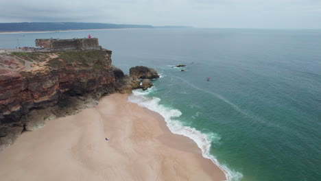 Aerial-view-over-the-beach-where-the-famous-Nazare-lighthouse-is-located-in-Portugal-on-a-summer-day