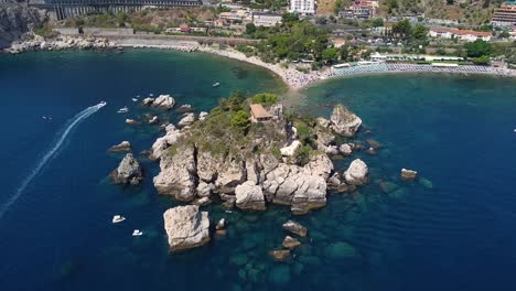 Aerial-drone-shot-zooming-into-Isola-Bella,-Taormina-in-Sicily,-with-boats-cruising-past,-blue-sea,-wide-view-of-the-bay-and-coastal-line-of-the-Italian-shore