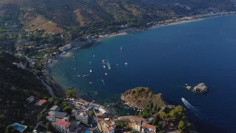 Toma-Aérea-Circular-Con-Dron-De-La-Costa-Italiana,-En-Taormina,-Sicilia,-Con-Barcos-Navegando-En-La-Bahía,-Mar-Azul-Y-Acantilados-Rocosos-Cubiertos-De-Hierba,-Apartamentos-Y-Complejo-Turístico-En-La-Colina