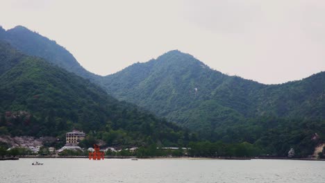 Ultrawide-view-of-Miyajima-Island's-Shinto-Shrine-at-high-tide-with-the-forested-mountains-of-Mount-Misen-in-the-background-on-a-hazy-overcast-day-in-Japan