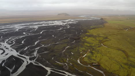 Luftaufnahme-Von-Gletscherflüssen,-Flussdelta,-Schwarzem-Vulkansand-Im-Süden-Islands,-Einer-Grünen-Ebene-Und-Einem-Berg-In-Der-Ferne,-Bewölkter-Tag,-Mündung,-Wunderschöner-Ort,-Wildnis,-Wasserlauf