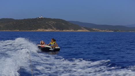 Zwei-Junge-Mädchen-Und-Frauen,-Die-Wassersport-Treiben,-Sitzen-Lachend-Auf-Einem-Donut,-Während-Sie-Von-Einem-Boot-Gezogen-Werden,-Das-über-Die-Wellen-Fährt,-Während-Sie-Am-Strand-Im-Sommerurlaub-Sind