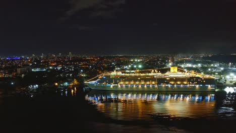 Kreuzfahrtschiff-Costa-Deliziosa-Nachts-Im-Hafen-Von-Sans-Souci-Mit-Beleuchteter-Stadt-Im-Hintergrund,-Santo-Domingo