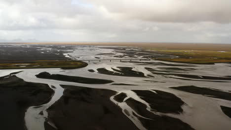 Aerial-view-of-river-delta,-black-volcanic-sand-in-South-Iceland,-cars-travelling-over-a-bridge,-glacial-rivers,-green-plain,-cloudy-day