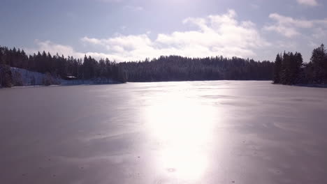 Aerial,-drone-shot-low-over-the-surface-on-a-icy-lake,-towards-forest,-sunny,-winter-day,-in-Risvann,-Birkeland,-Agder,-South-Norway