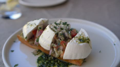 Slow-motion-footage-rotating-around-a-plate-of-bruschetta-topped-with-Gozo-cheese,-chopped-tomatoes,-onions-and-pesto