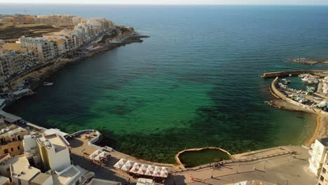 360-degree-aerial-footage-revealing-the-harbor-of-Marsalforn,-buildings-and-sea-front-during-golden-hour-at-sunset-in-Gozo,-Malta