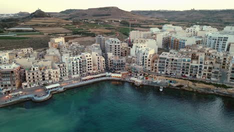 Aerial-footage-showing-the-harbor-of-Marsalforn,-buildings-and-sea-front-front-during-golden-hour-at-sunset-in-Gozo,-Malta