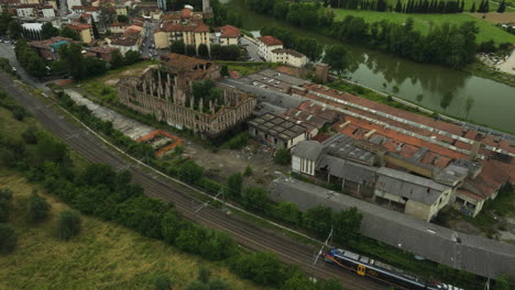 Tren-Sobre-Vía-Férrea-Que-Pasa-Por-Una-Fábrica-De-Cerámica-Abandonada-Junto-Al-Río-Arno-En-Le-Sieci,-Italia