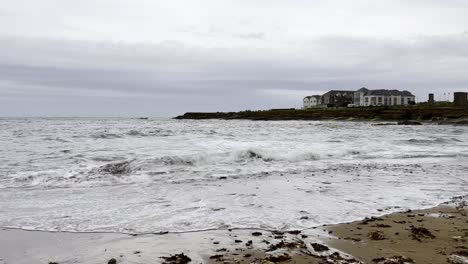 Olas-En-La-Playa-De-Spanish-Point,-Irlanda.-Las-Olas-Golpean-Las-Rocas-De-La-Playa-Y-Se-Puede-Ver-El-Hotel-A-Lo-Lejos.