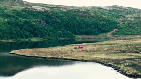 Un-Hombre-Y-Su-Perro-Se-Relajan-Junto-A-Su-Tienda-De-Campaña-En-La-Orilla-Del-Lago-Interior-Kjøåkertjønna-En-Osen,-Trøndelag,-Noruega---Toma-Aérea-Con-Dron