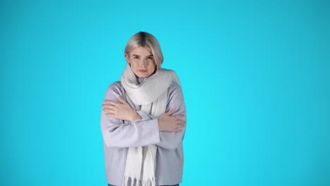 Porträt-Einer-Jungen-Blonden-Frau,-Die-Friert,-Trägt-Schal-Und-Grauen-Pullover,-Studioaufnahme-Mit-Blauem-Hintergrund