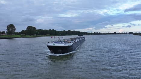 Großes-Frachtschiff,-Das-Auf-Einem-Breiten-Fluss-Mit-Grünen-Ufern-Und-Bewölktem-Himmel-Im-Hintergrund-Navigiert