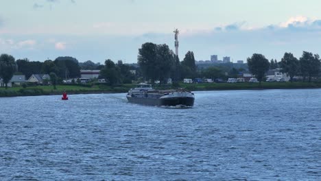 Cargo-ship-sailing-on-a-river-with-a-village-and-trees-in-the-background,-overcast-day