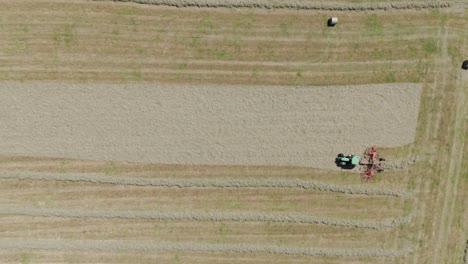 Top-down-aerial-view-of-a-green-tractor-milling-rows-of-freshly-cut-grass,-preparing-it-for-bailing,-in-a-vast,-neatly-patterned-agricultural-field