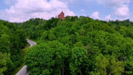 Drone-reveals-Turaida-castle,-Latvian-woodlands,-road-through-green-rural-area