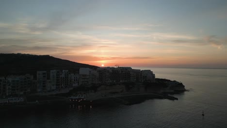 Aerial-footage-revealing-the-sun-setting-behind-the-harbor-of-Marsalforn,-buildings-and-sea-front-front-in-Gozo,-Malta