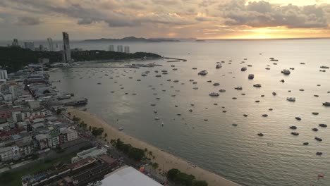 Aerial-trucking-pan-above-Pattaya-city-and-beach-as-ocean-waves-crash-on-sandy-shores-with-boats-anchored-off-shore-at-sunset