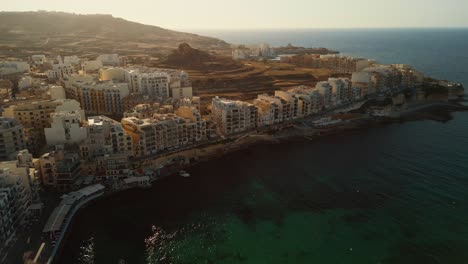 18-degree-aerial-footage-showing-the-harbor-of-Marsalforn,-buildings-and-sea-front-front-during-golden-hour-at-sunset-in-Gozo,-Malta