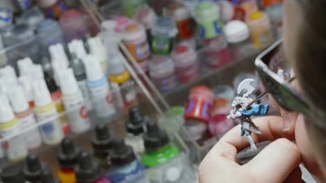 Artist-with-Glasses-Painting-Miniature-Fantasy-Figure-Model-Creature-with-Vast-Array-of-Paint-Color-Pots-for-Creative-Choices-in-Background-4K