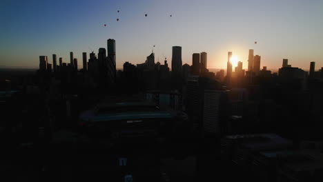 AERIAL:-Hot-air-balloons-above-the-silhouette-Melbourne-skyline,-during-sunset