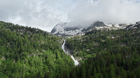 Cascading-waterfall-amidst-lush-green-forest-and-snowy-mountain-peaks-under-a-cloudy-sky