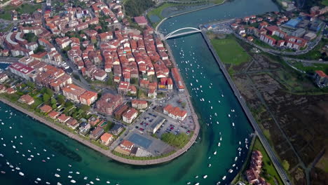 Aerial-view-of-a-charming-coastal-town-Santurzi,-Bilbao-with-tightly-packed-red-roofed-buildings,-winding-streets,-a-river-lined-with-boats,-bridge-connects-the-town,-surrounded-by-greenery-and-water