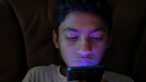 Teenage-boy-sitting-on-sofa-using-smart-phone-at-night