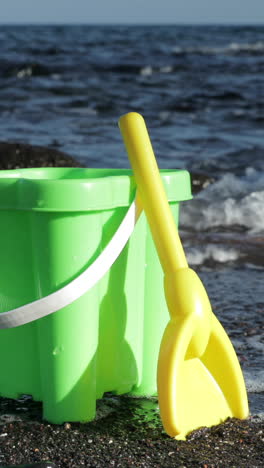 Plastikeimer-Und-Schaufel-Auf-Einem-Schwarzen-Sandstrand-Mit-Dem-Meer-In-Vertikaler