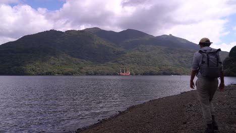Male-Hiker-walking-into-frame-at-Hakone-Lake,-waving-at-famous-Pirate-Ship-in-distance
