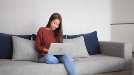 Asian-woman-model-sitting-on-the-grey-cozy-sofa-with-the-laptop-on-laps-and-typing-on-a-notebook-with-a-happy-face-expression,-front-view-slow-motion-handheld