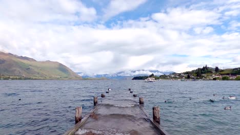 Peaceful-view-across-wanaka-lake-with-submerged-pier,-ducks,-boats-and-mountains-in-the-scene