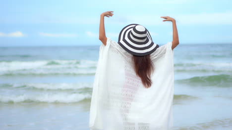 A-young-woman-in-beach-clothes-and-a-big-hat-walks-on-the-sandy-beach-with-arms-outstretched-on-the-beach-and-enjoys-the-view-of-the-sea