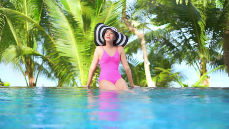 Young-female-tourist-sitting-on-the-edge-of-a-swimming-pool-wearing-a-pink-one-piece-swimwear-and-a-floppy-hat-with-palm-trees-blurred-in-the-background