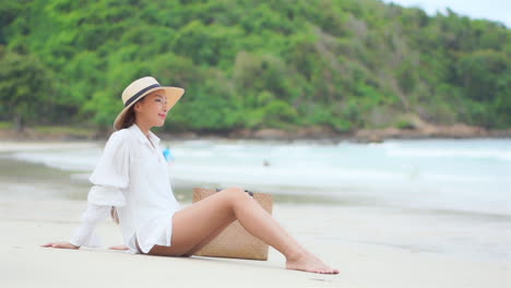 Asian-woman-sitting-on-a-white-sand-beach-in-front-of-the-sea-wearing-a-white-blouse-shirt-and-straw-hat,-tropical-island-nature-in-the-background,-slow-motion-side-view-handheld