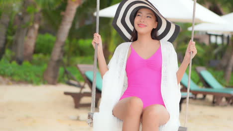 Asian-tourist-swinging-at-the-beach-in-a-tropical-destination-during-sunny-day-wearing-pink-swimsuit,-bohemian-transparent-cloth-and-a-big-floppy-hat