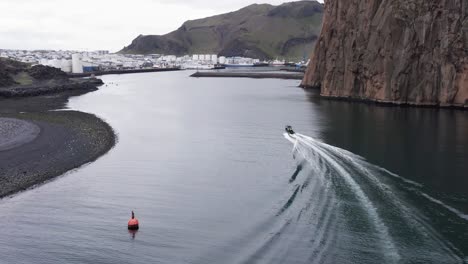 Rigid-hull-inflatable-speed-boat-entering-inlet-of-Vestmannaeyjar,-follow-aerial