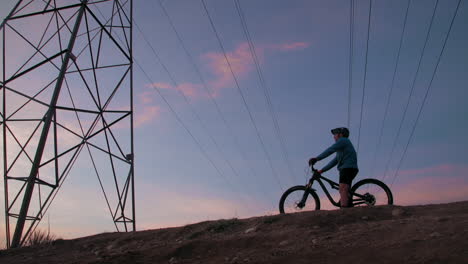 A-male-mountain-biker,-pulls-under-a-power-line-tower-to-observe-a-dramatic-setting,-as-the-low-sun-silhouettes-his-form