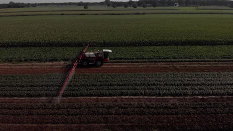 Sideways-aerial-follow-of-a-tractor-dragging-wide-spraying-arms-and-stock-of-liquid-in-farmland-surroundings-with-various-crops-in-the-background-and-foreground