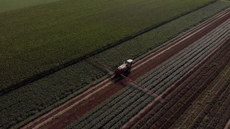 Aerial-following-and-pan-around-behind-a-tractor-dragging-wide-spraying-arms-and-stock-of-liquid-moving-along-straight-vegetation-line