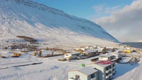 Seguimiento-Aéreo-Sobre-Un-Pequeño-Pueblo-Nevado-De-Invierno-Al-Pie-De-Una-Colina-Montañosa-Cubierta-De-Nieve-En-Los-Fiordos-Del-Oeste-En-Islandia