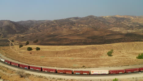 A-train-hauls-cargo-beneath-mountains-and-hills-burned-black-by-wildfires-near-Caliente,-California---aerial-view