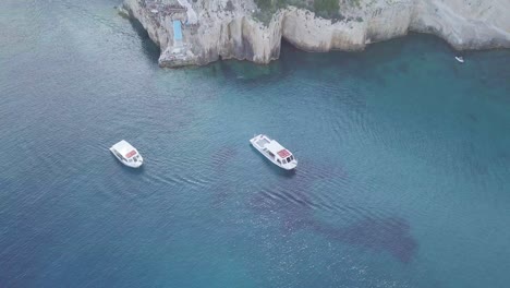 Drone-footage-of-cliffs-on-the-coast-of-Zakynthos-Greece,-featuring-two-yachts,-blue-seas-and-several-buildings-on-top-of-the-cliffs