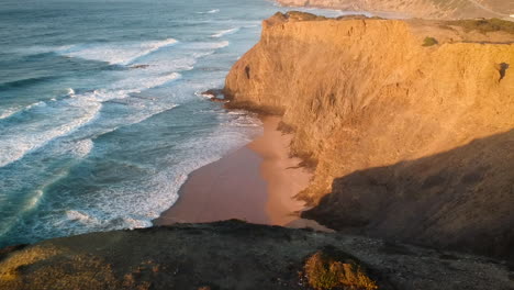 Aerial-drone-shot-of-waves-hitting-a-beach-with-cliffs-during-a-sunset-in-southern-Portugal